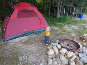 Toddler camping, standing in front of red tent holding a stick to an empty fire pit