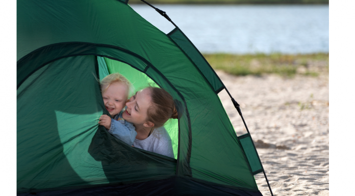 Mother and baby camping, peeking out of a green tent on a sandy shore