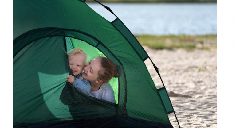 Mother and baby camping, peeking out of a green tent on a sandy shore