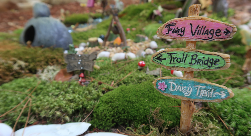A sign that states the directions to the fairy village, troll bridge, and daisy trails with the fairy village blanketed with moss in the background