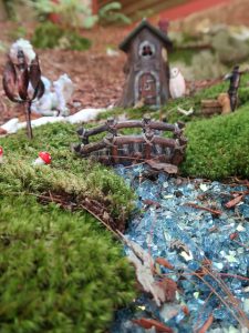 A view of a miniature bridge over a blue glass stream in front of a small fairy cottage.
