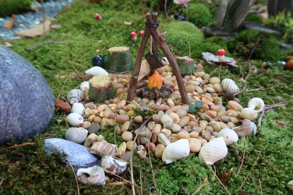 A miniature fire ring with a stove getting warmed up by the raging fire. There are tree stumps surrounding the fire ring for fairies to sit on.