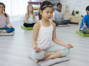 Child in front of 4 other children, sitting cross-legged with eyes closed and hands facing upward, practicing breathing exercises for kids