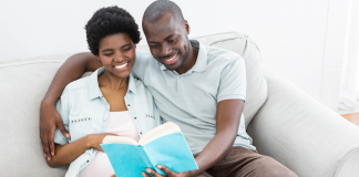 Two people sitting on a couch. One person has their arm around a pregnant person and they are reading a book together. Books to read when you're pregnant.