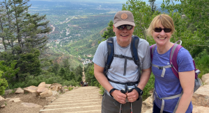 A man and woman at the top of the Manitou Incline stairs, Manitou Springs, Colorado. They are standing, smiling, wearing backpacks.
